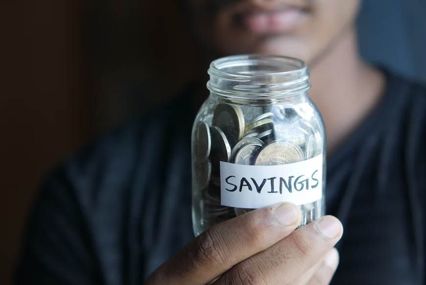A man holding a jar full of coins in front of him with a label "SAVINGS".