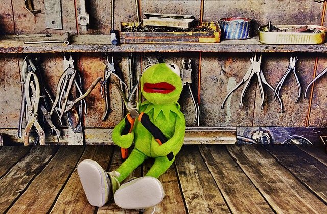 A green stuff toy that looks like a frog sitting down a wooden platform while holding a pliers with a few more tools behind him.