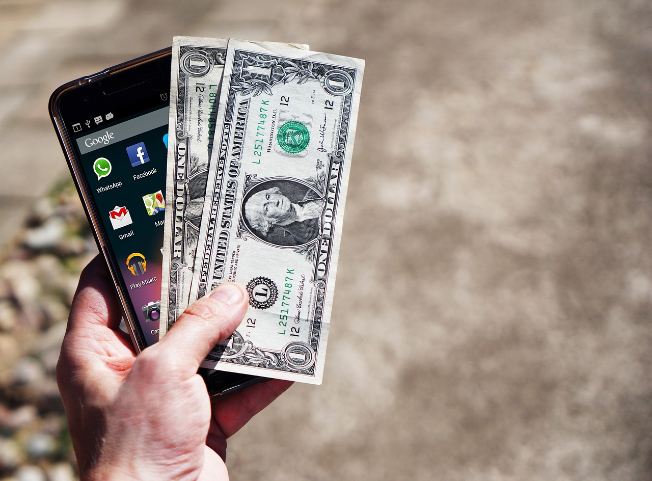Man holding a mobile phone with 2 dollar bills on top.