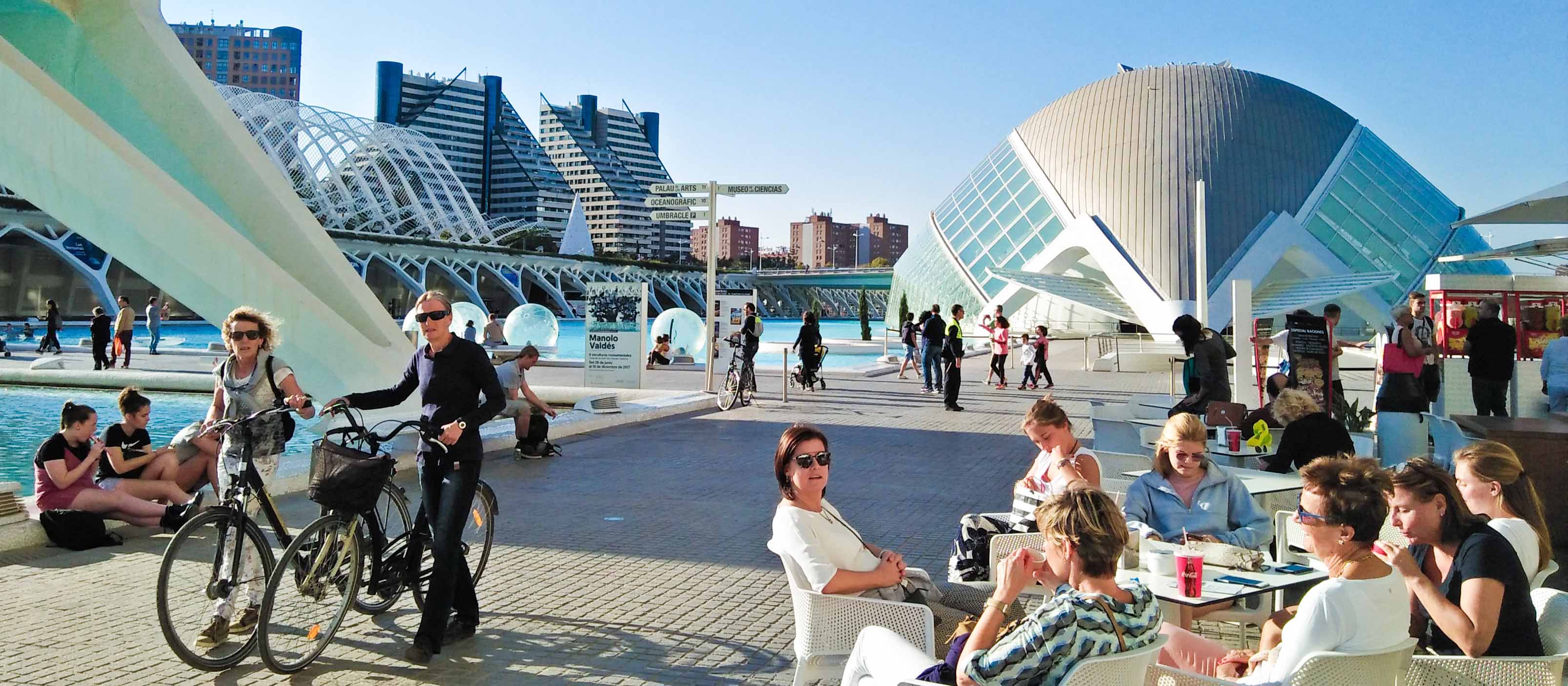 Tourists enjoying the summer by dining outside, walking their bicycles, sitting near the pool, and chatting with everyone else in Ciudad de las Artes y las Ciencias, Valencia, Spain.