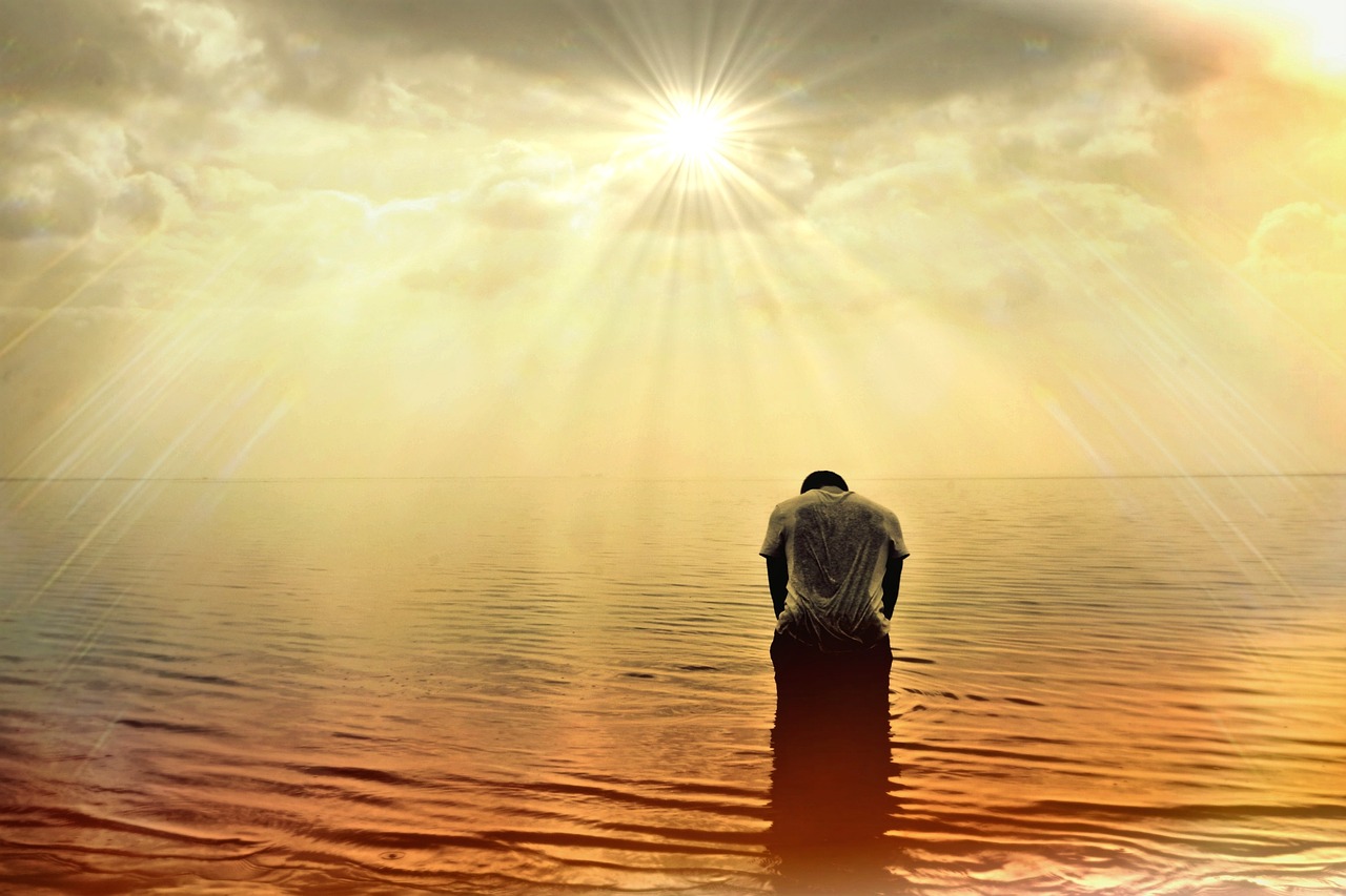 A man in the shallow beach bowing his head while facing the sun as if reciting a prayer.