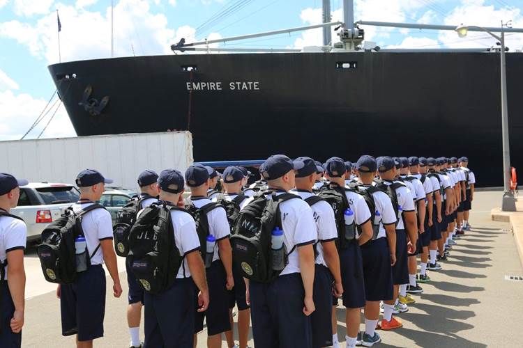 Cadets wearing uniforms of white t-shirt, navy blue shorts and caps, and backpack in formation near a huge cargo vessel.