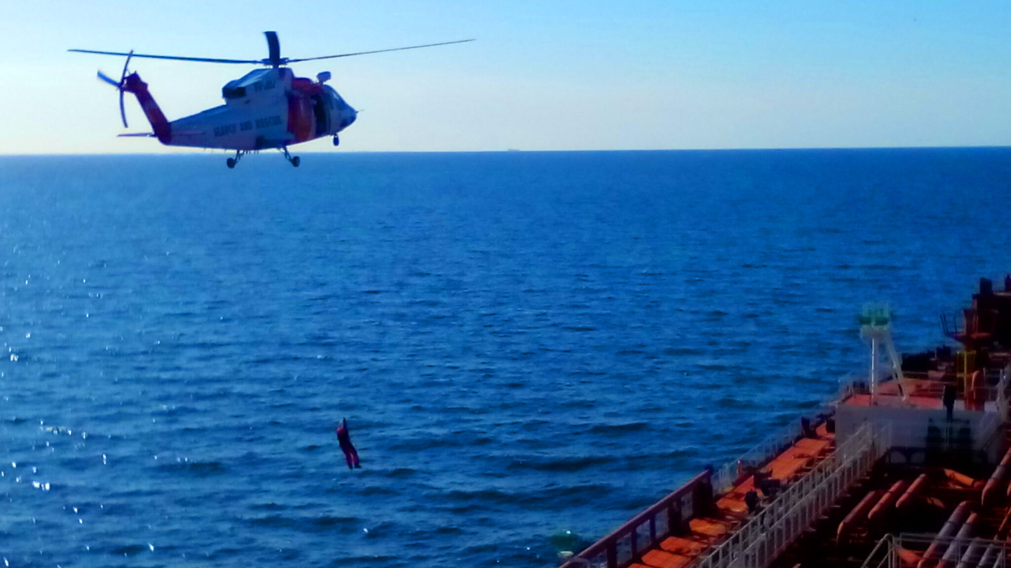 A rescuer hanging down the winch as the helicopter approaches to the tanker vessel's main deck.