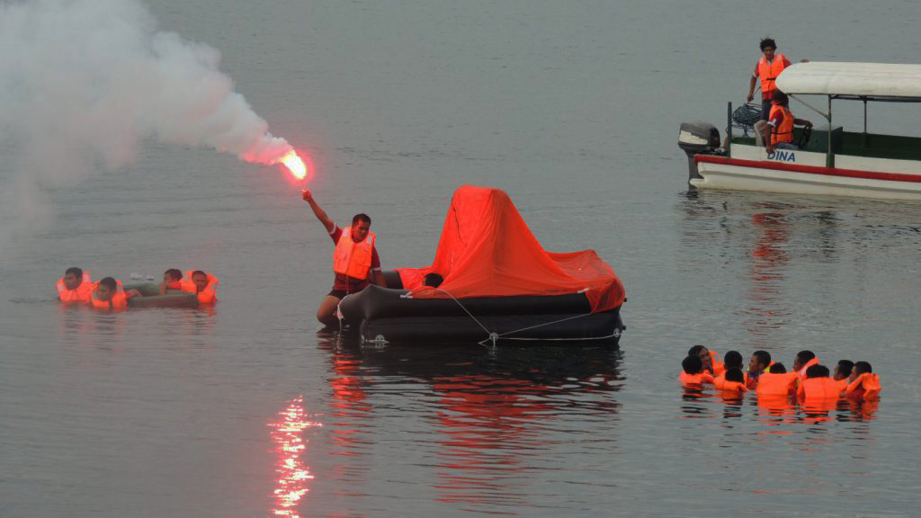 Basic Training Practical Exercise with life raft at sea. Trainees are in the water performing personal survival techniques while another crew is on board the liferaft holding a hand flare.