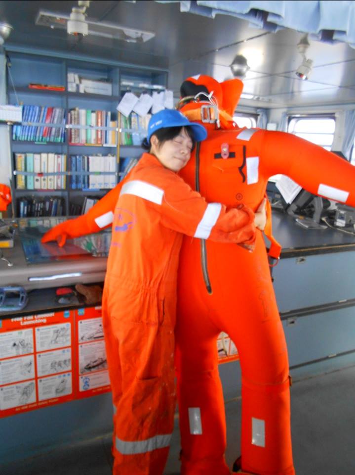 Hiroka wearing a cap and an orange coverall hugging an inflated immersion suit during her maintenance inspection.