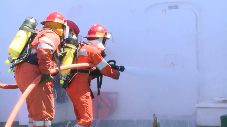 Fire fighters with firehose and nozzles extinguishing flames on board ship.