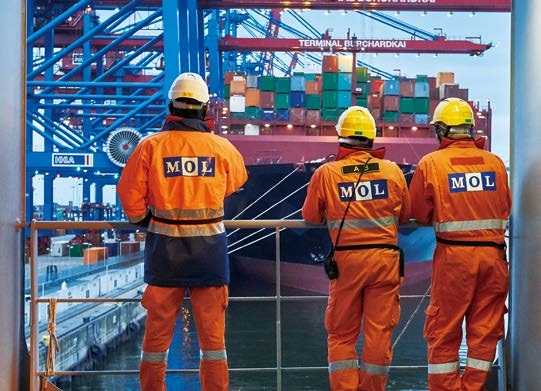 Crew of Mitsui OSK Lines in standing near the railings of their vessel wearing full PPE: coverall, helmet and handheld radios.