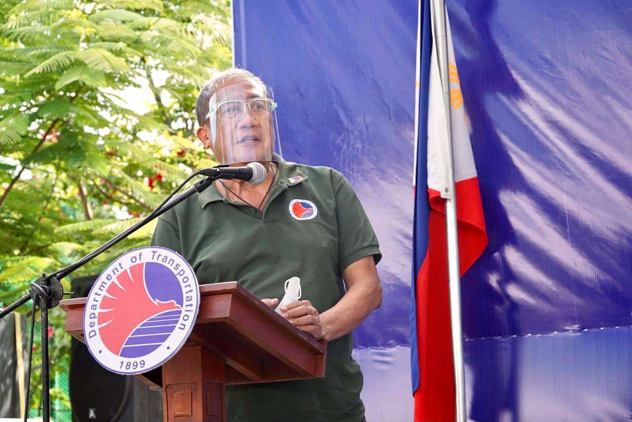 Secretary Art Tugade in a speech wearing face shield and the Philippine flag behind him.