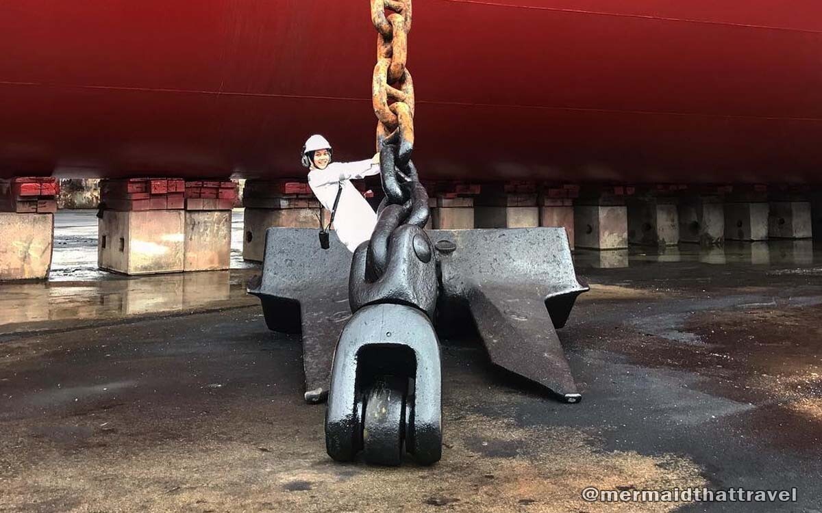 A ship's anchor lowered down in a shipyard while a crew stands on top of it holding the huge chain.