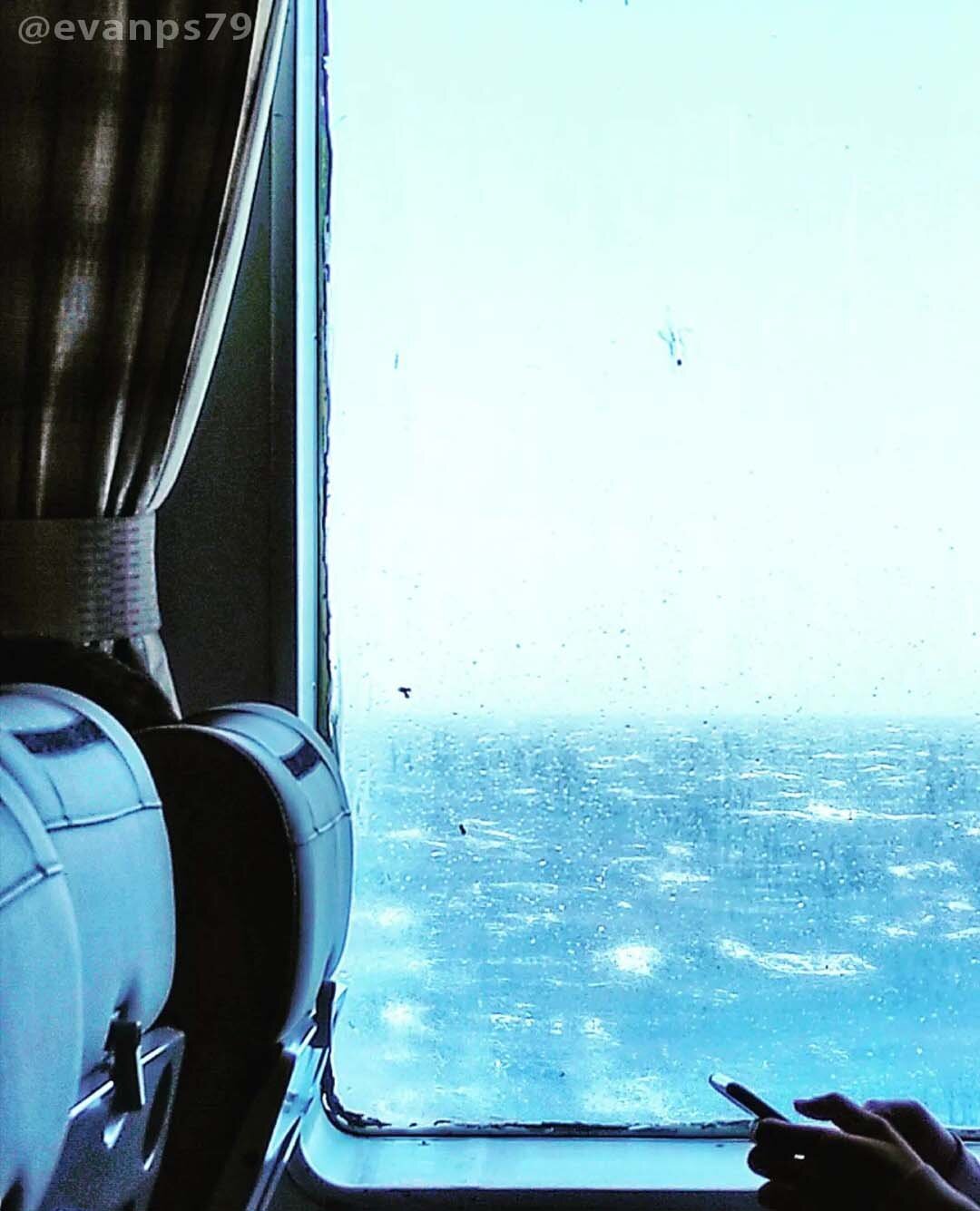 A person using his phone near the ship's porthole showing the rainy environment outside.
