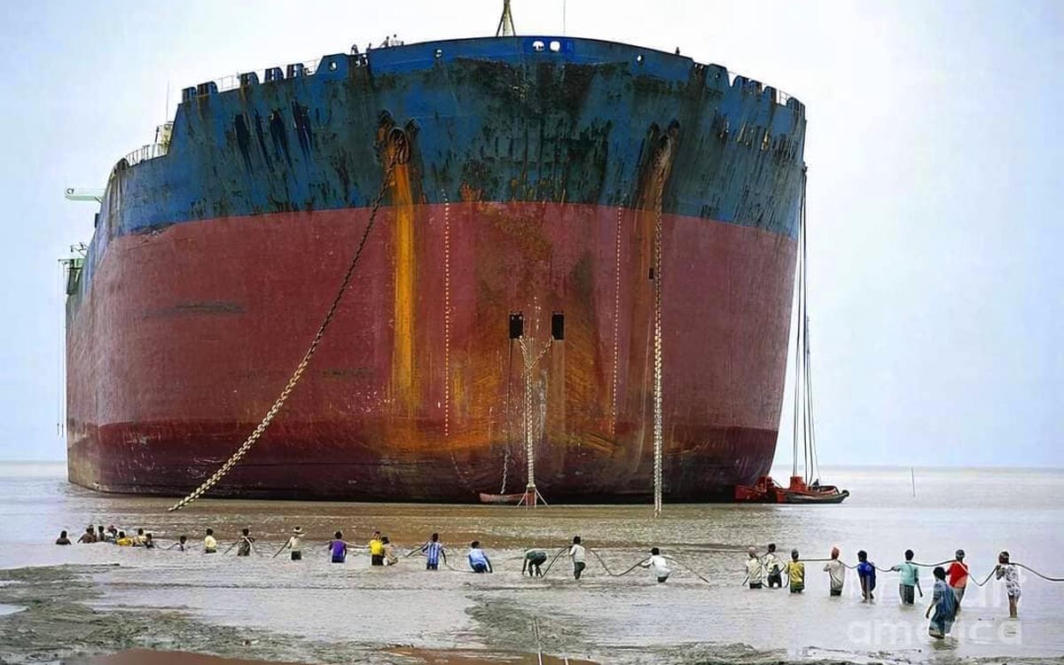 A huge merchant ship beached near the shore and people are approaching it.