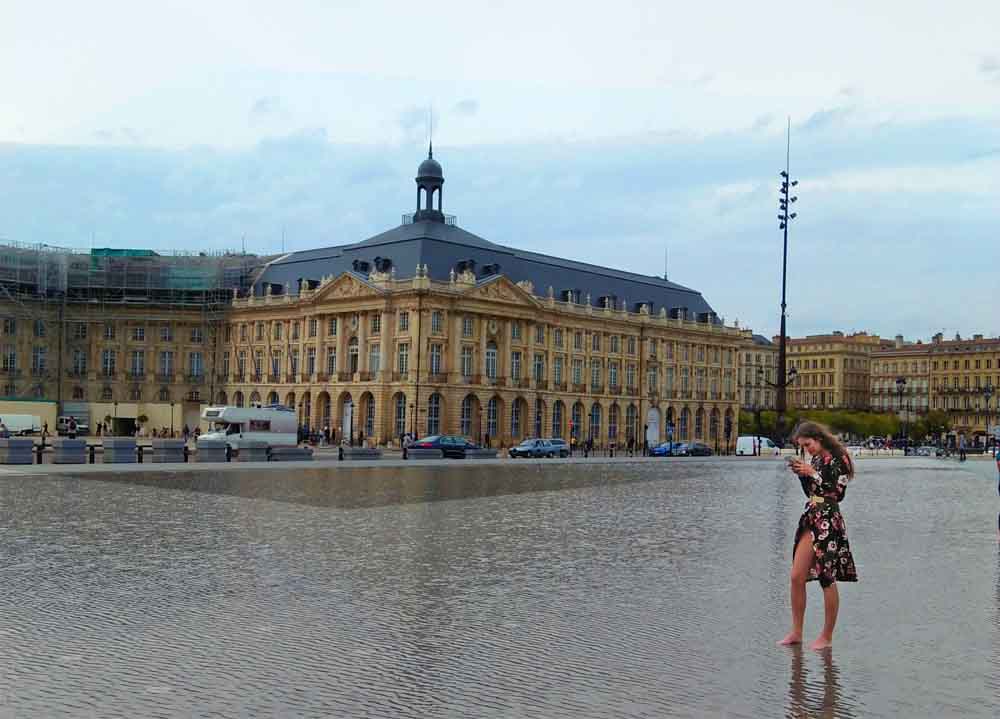 A woman standing in a pool of water  while barefoot in front of the Palais de la Bourse in Bordeaux, France. The building is reflected in the water, and the woman is looking at the camera.