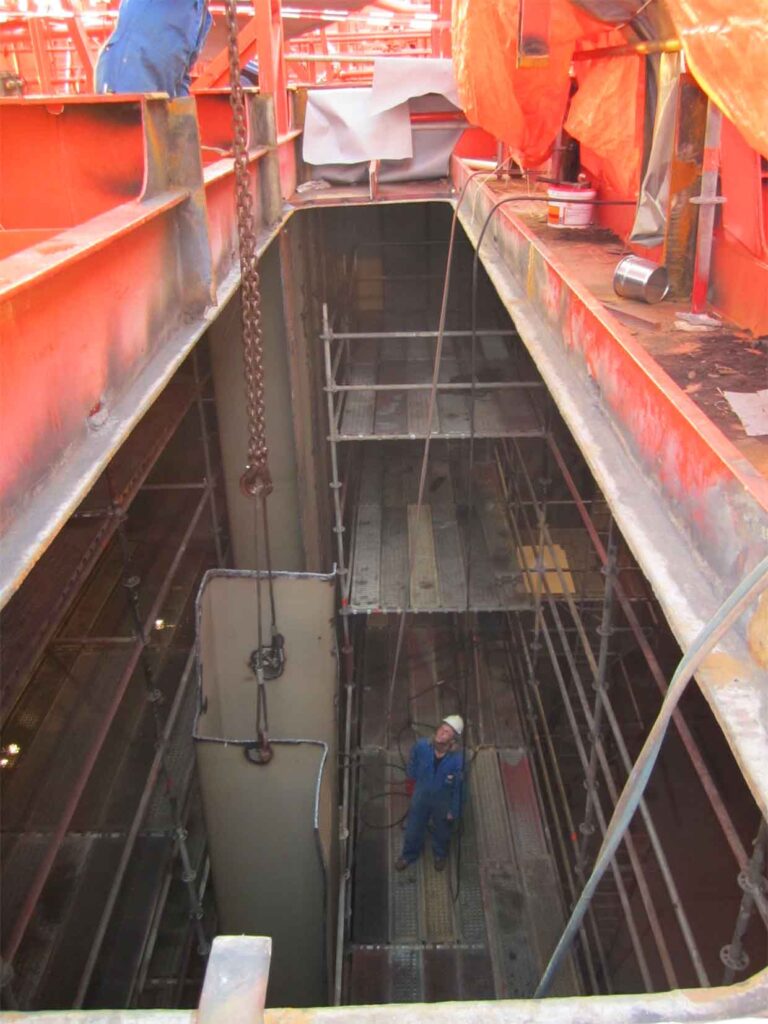 A yard worker assisting in heaving up a damaged corrugated bulkhead of a tanker vessel inside a cargo tank while its deck is ripped open. This repair is covered by marine insurance.