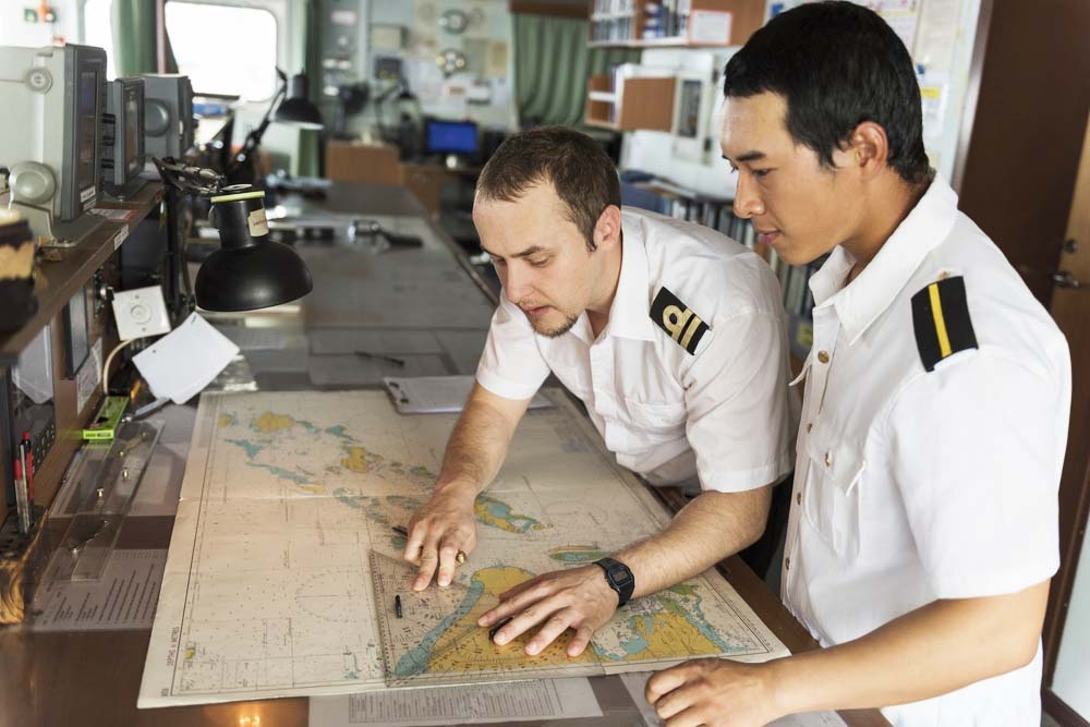 A Second Officer teaching a young deck cadet about position fixing on a nautical chart.