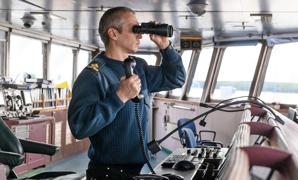 A ship's Third Officer looking through the binocular while holding a VHF radio in his right hand.