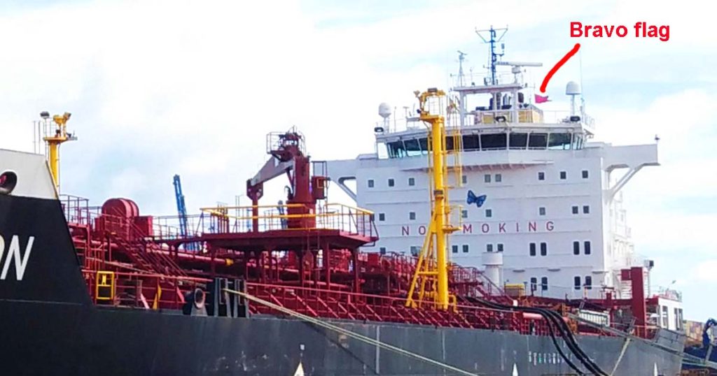 A tanker ship in port performing cargo operation and displaying the red Bravo Flag on its mast.